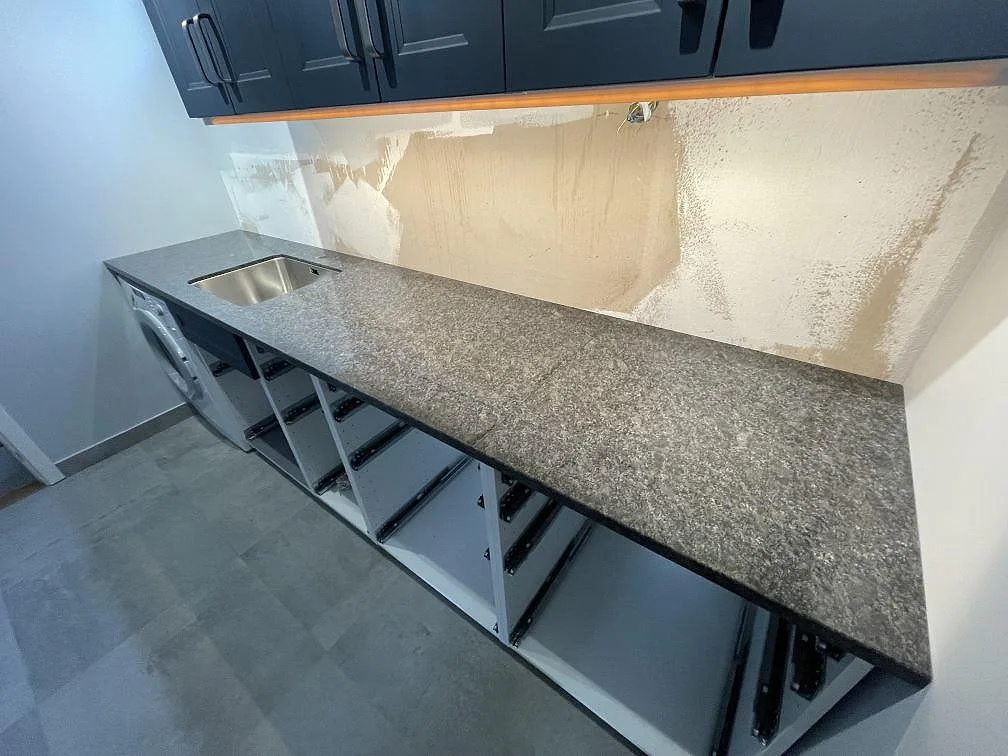 Lång polerad bänkskiva i Steel Grey granit med jämn spräcklig yta och öppna underskåpsstommar i Tumba.