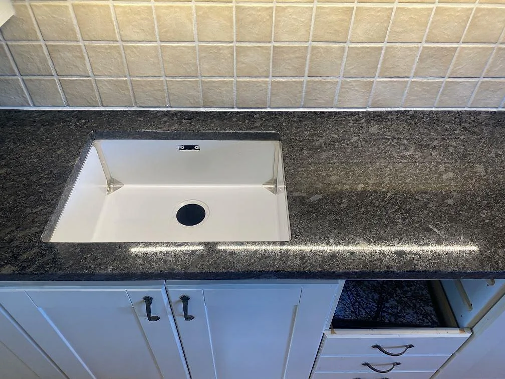 Underlimmad vit diskho i polerad Steel Grey granitbänkskiva mot ljust kaklat stänkskydd i ett kök i Vejbystrand.
