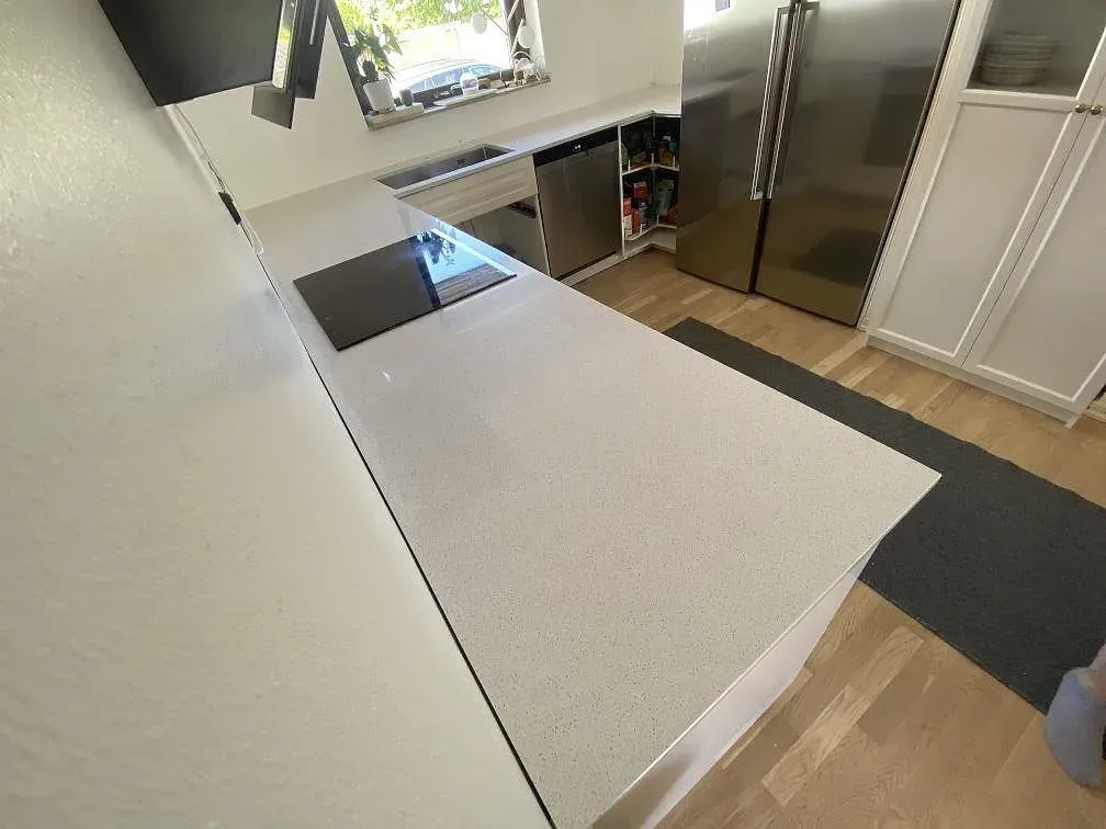 Köksbänkskiva i vit Technistone Crystal Quartz White, polerad, som löper runt ett kök med infälld induktionshäll och diskmaskin i Malmö.