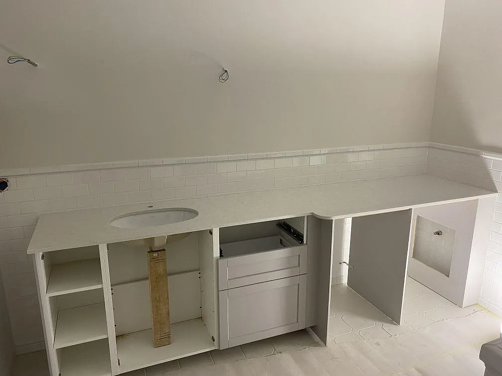 Bänkskiva i polerad Technistone Glencoe med vitt tvättställ, vit kakelvägg och öppna stommar under montering i Ängelholm.