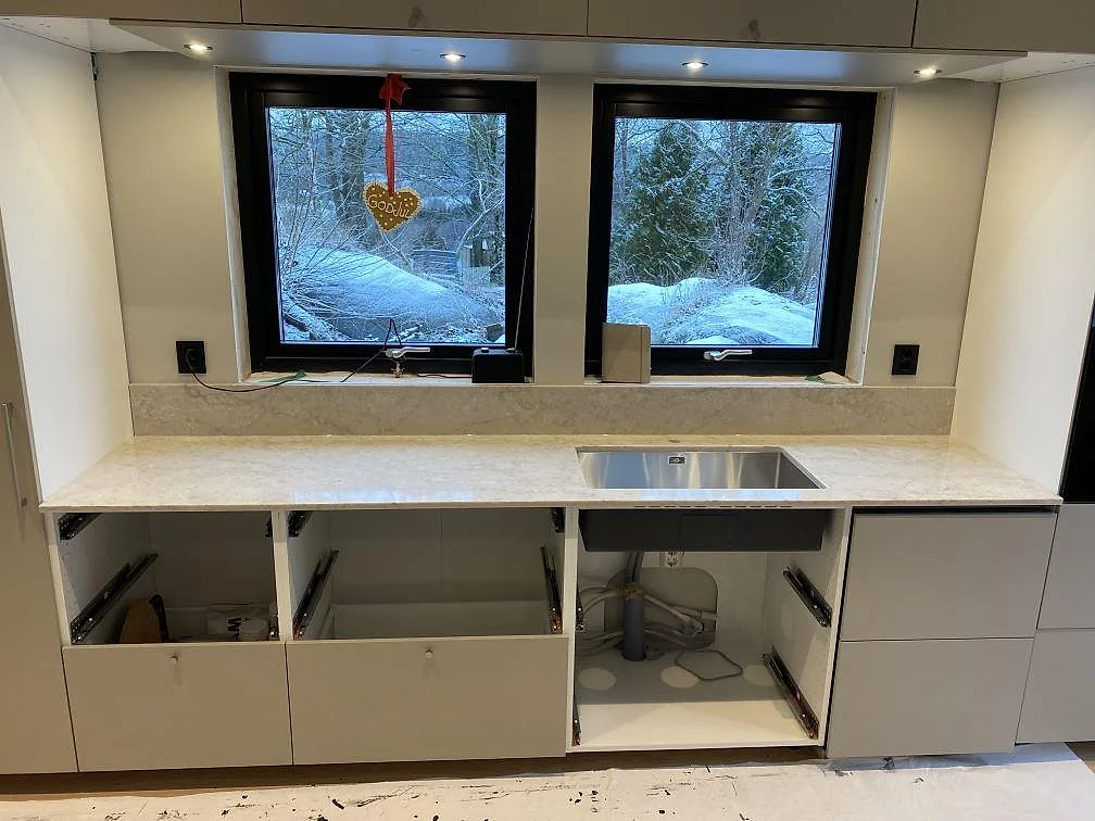 Polerad köksbänkskiva i Technistone Glencoe med uppvikt stänkkant och rostfri diskho vid fönster i Alafors.