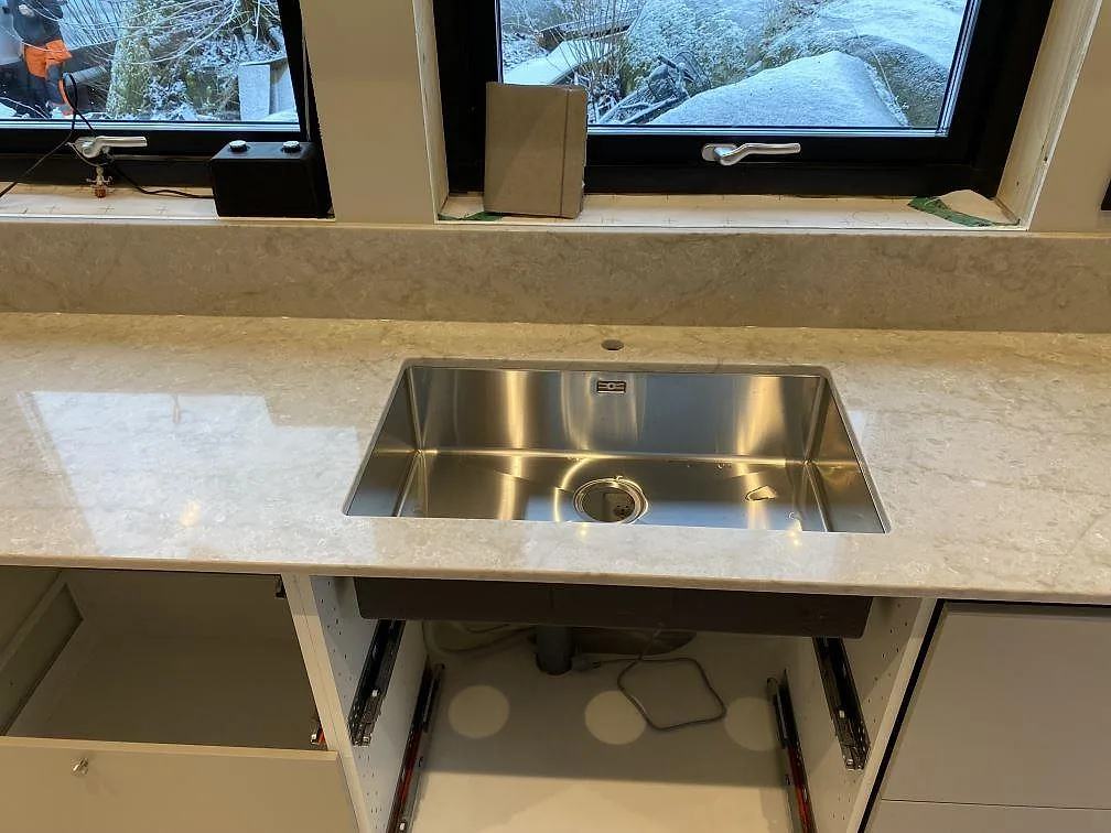 Rostfri diskho i bänkskiva av polerad Technistone Glencoe med låg stänkkant under fönster i Alafors.