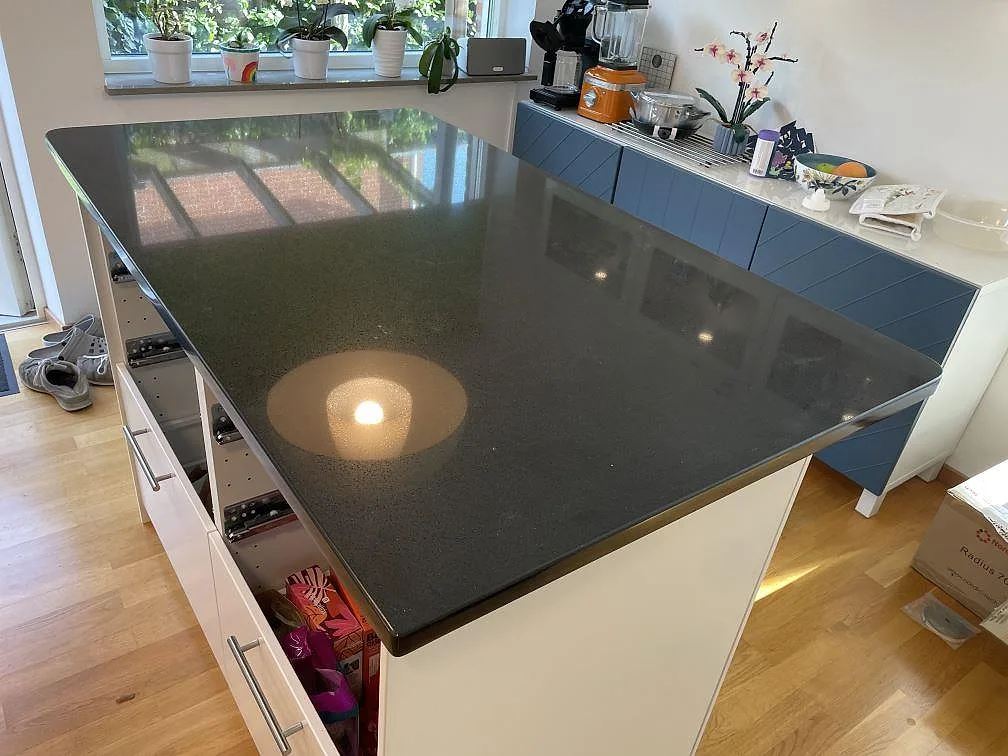 Stor köksö i mörk polerad Technistone Gobi Black komposit med trägolv och blå förvaringsmöbel i Dalby.