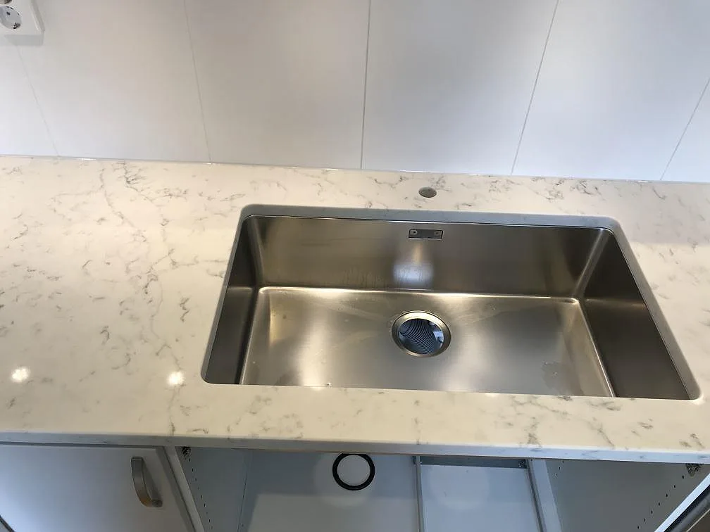 Underlimmad rostfri diskho i en köksbänkskiva av Technistone Mystery White i Åhus, med separat borrat blandarhål i den polerade stenen.