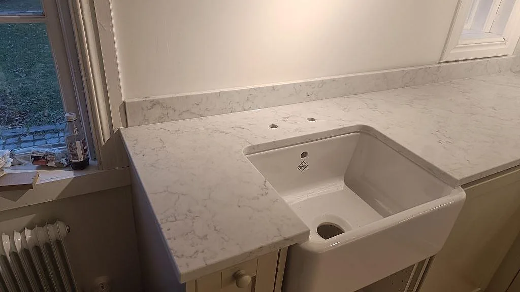 Närbild av polerad Technistone Mystery White som köksbänkskiva med vit keramisk farmhouse-diskho och borrhål för blandare i Åhus.