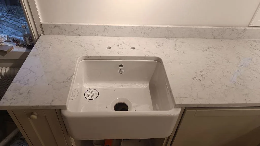 Topvy av polerad Technistone Mystery White runt farmhouse-diskhon, med grå ådring och uppvikt stänkkant i köket i Åhus.