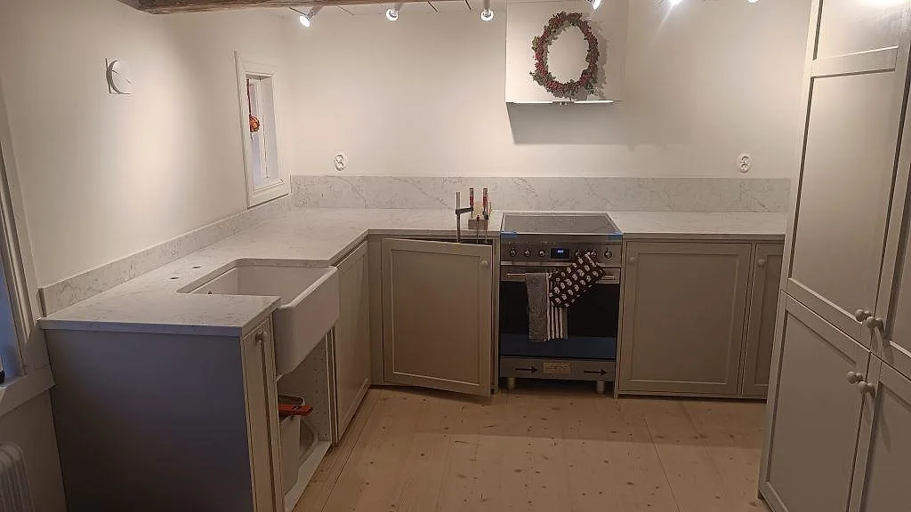 Köksbänkskiva i polerad Technistone Mystery White med låg stänkkant, vit keramisk farmhouse-diskho och spis i ett kök i Åhus.