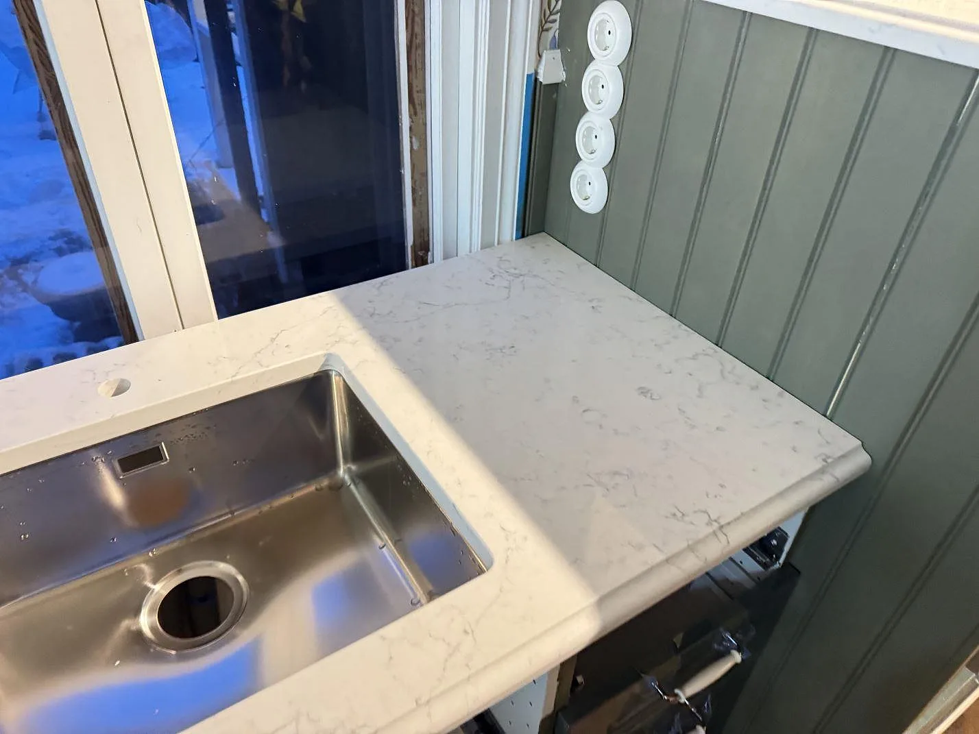Rostfri underlimmad diskho och uppvikt stänkkant i polerad Technistone Mystery White mot grön pärlspont i ett kök i Båstad.