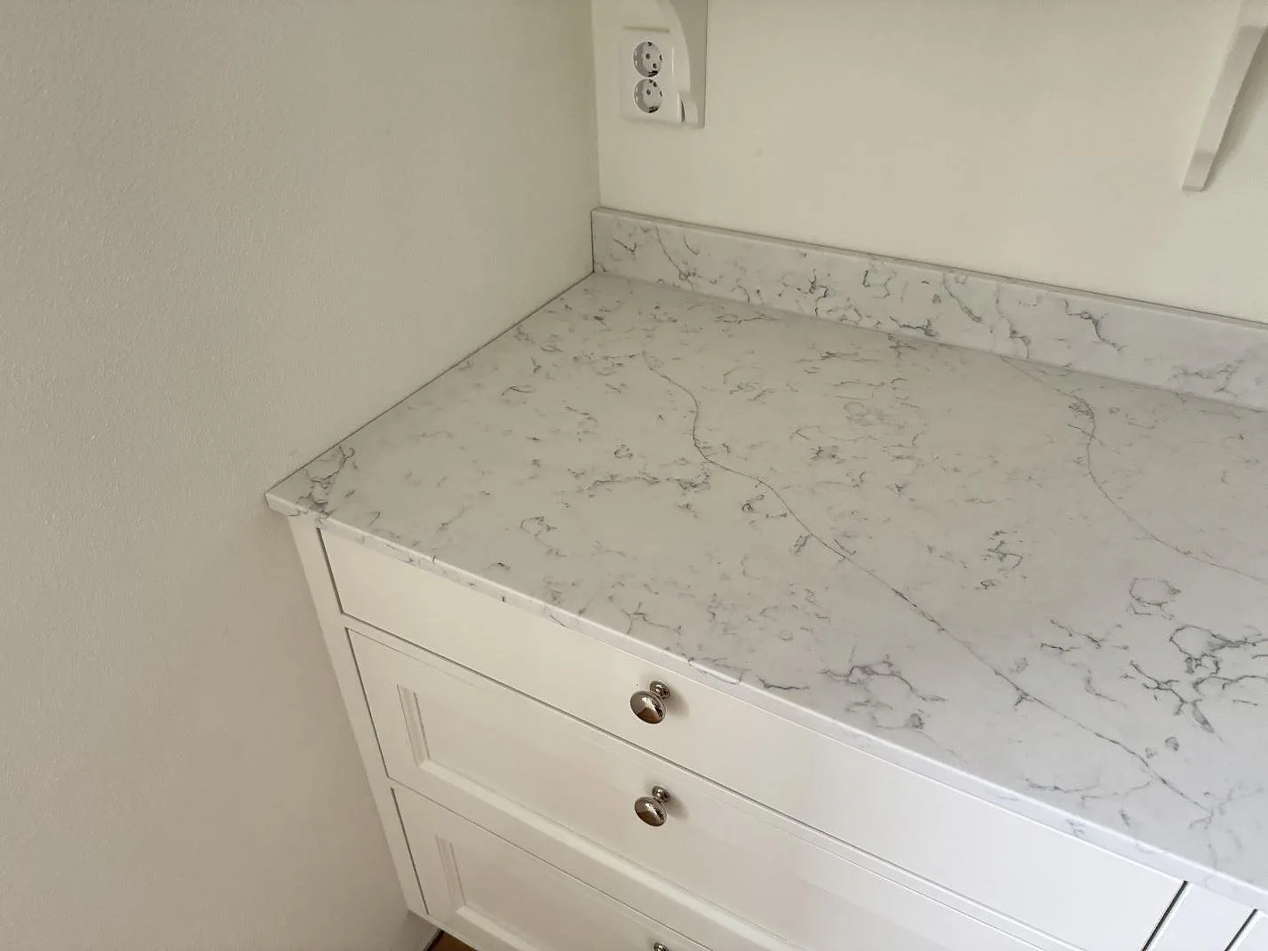 Vänster del av köksbänkskiva i polerad Technistone Mystery White med låg stänkkant över vita köksluckor i Barsebäck.