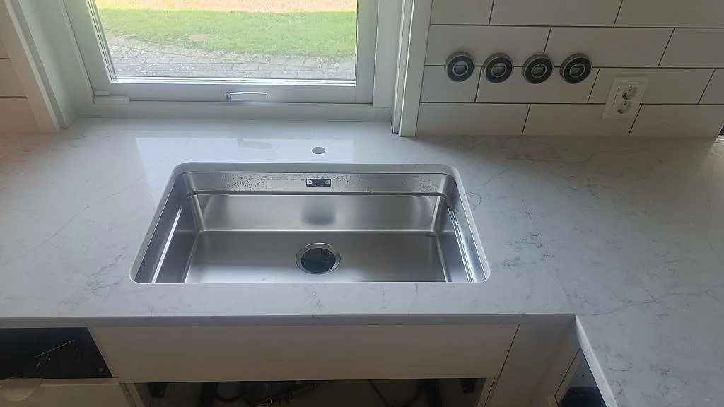 Närbild på rostfri diskho monterad under polerad Technistone Mystery White med ljus ådring i Bunkeflostrand.