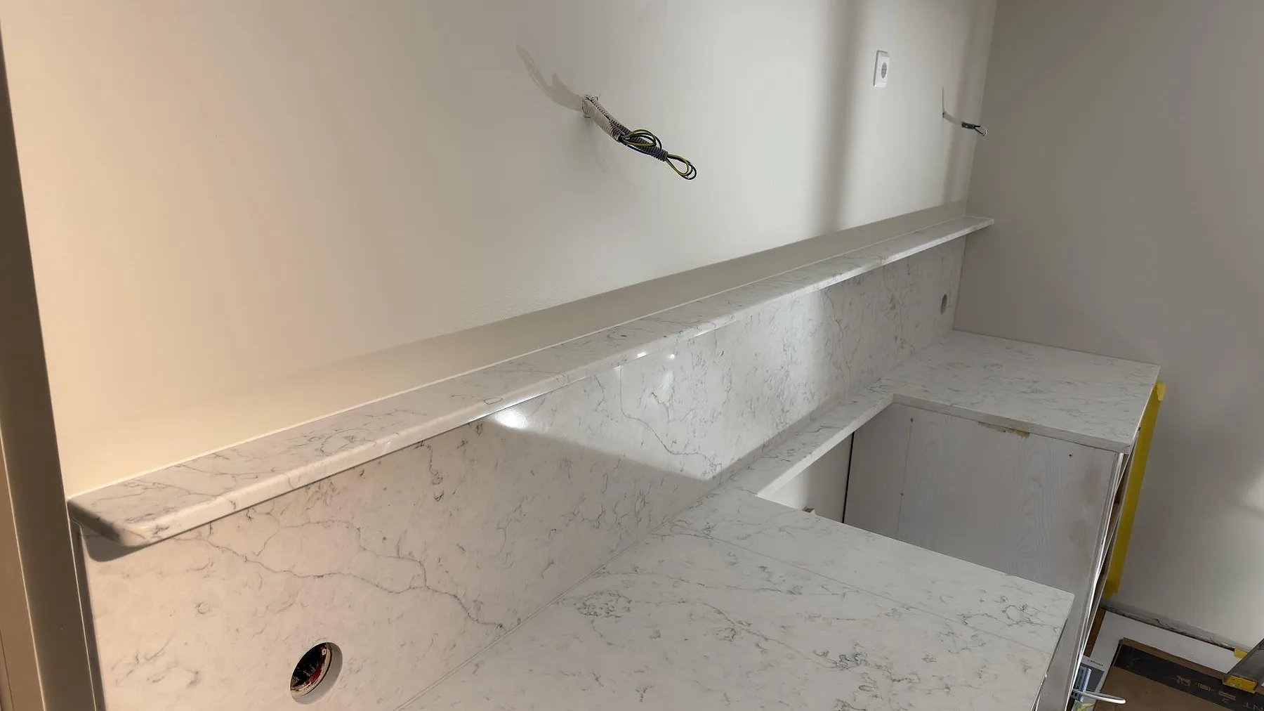 Polerad Technistone Mystery White i kök med uppvikt stänkskydd, smal stenhylla och förberett urtag under montering i Lidingö.