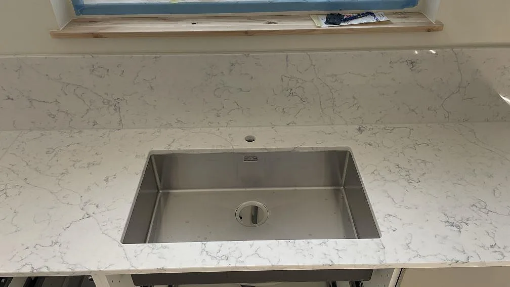Rostfri diskho i en polerad köksbänkskiva av Technistone Mystery White med stänkskydd under fönster i Lomma.