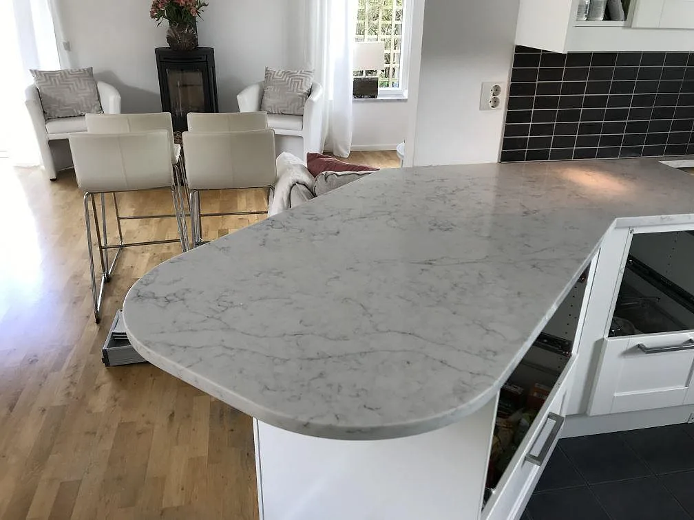 Svängd köksö i polerad Technistone Mystery White med diskret grå ådring, sedd mot vardagsrummet i ett kök i Rydebäck.