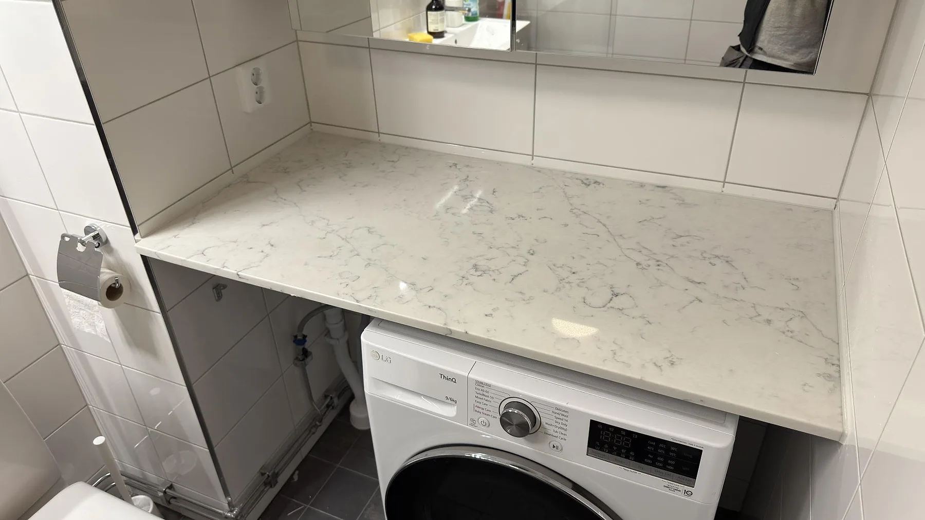 Bänkskiva i polerad Technistone Mystery White ovanför tvättmaskin i ett kaklat badrum i Stockholm, med ljus ådring i kvartskompositen.