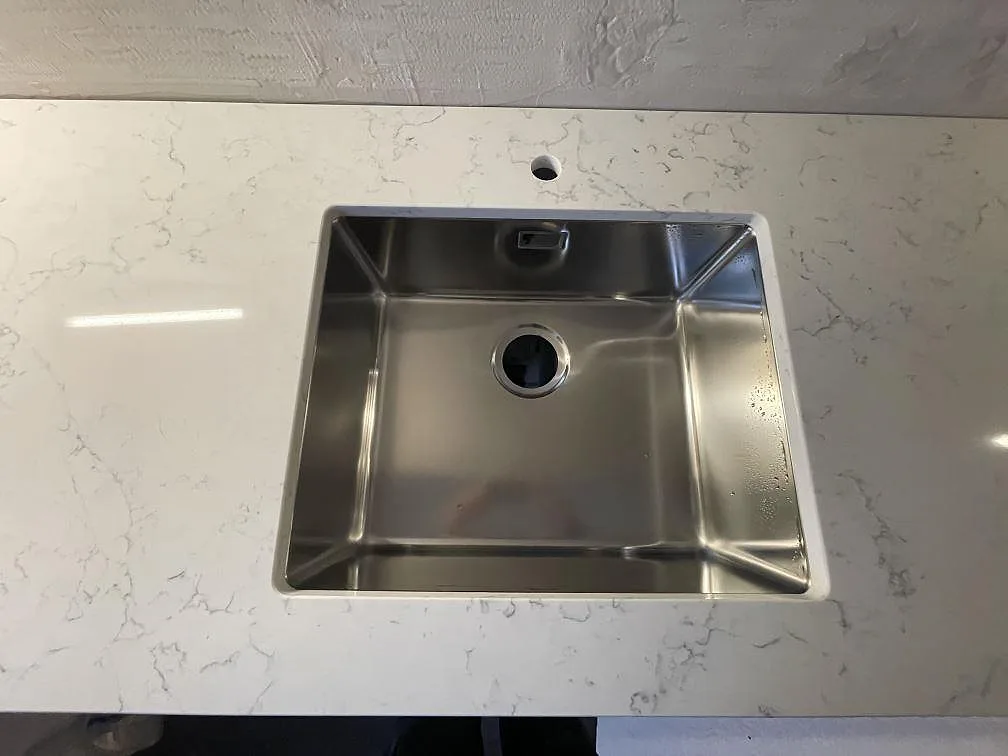 Underlimmad rostfri diskho med borrat blandarhål i en polerad bänkskiva av Technistone Mystery White i ett kök i Stockholm.