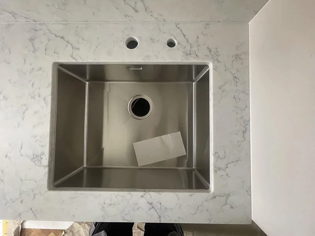 Underlimmad rostfri diskho infälld i köksbänkskiva av Technistone Mystery White, med två förborrade blandarhål och polerad stenyta, i ett kök i Stockholm.