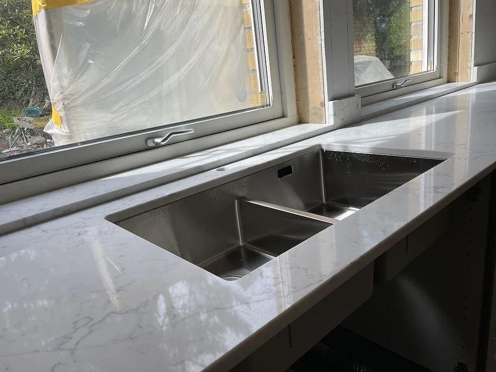 Närbild på polerad Technistone Mystery White köksbänkskiva med dubbel diskho vid fönster i ett kök i Tyresö.