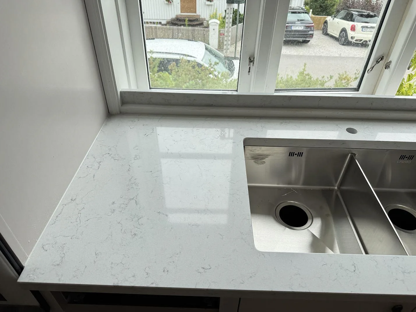 Polerad köksbänk i Technistone Noble Carrara med rostfri dubbelho under fönster i ett kök i Båstad.