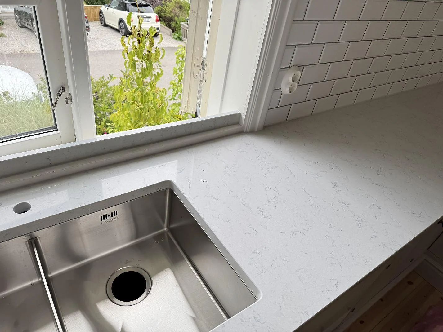 Köksbänkskiva i polerad vit kvartskomposit Technistone Noble Carrara med diskho, fönster och arbetsyta mot kakel i Båstad.
