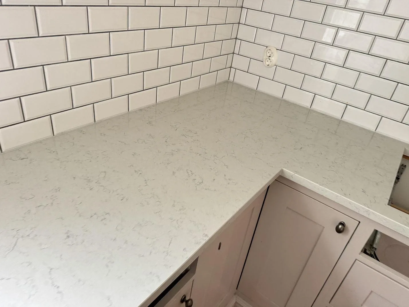 L-formad köksbänk i polerad Technistone Noble Carrara med vit kakelvägg och ljusa luckor i Båstad.