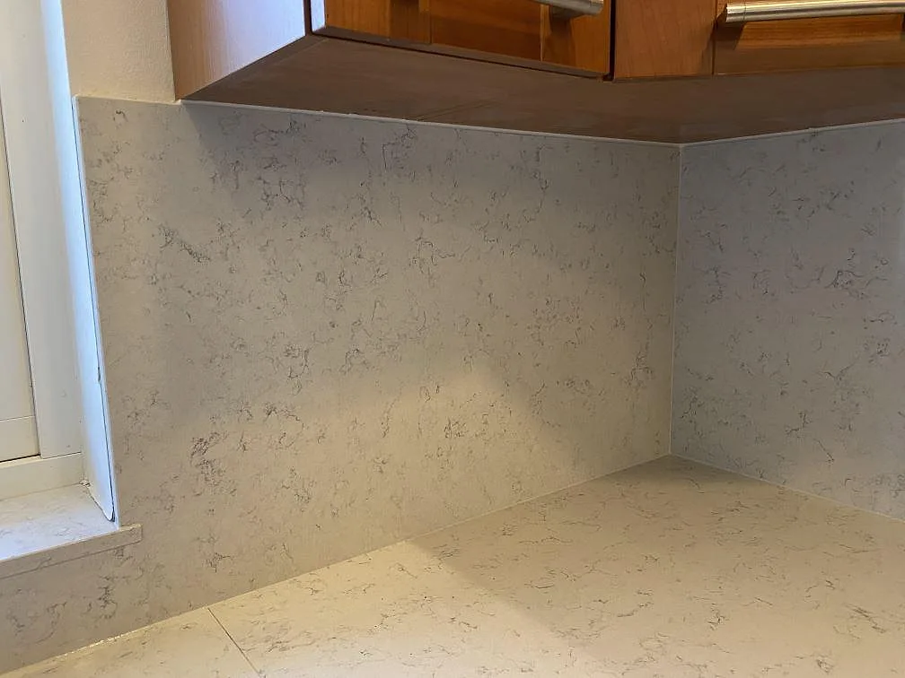 Hörn av köksbänkskiva och stänkskydd i polerad Technistone Noble Carrara vid fönster och träskåp i Blentarp.