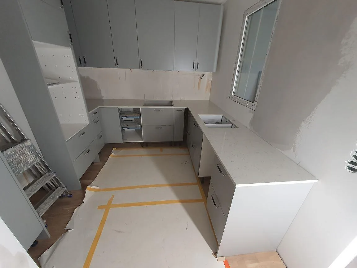 Köksbänkskiva i vit Technistone Noble Carrara, polerad kvartskomposit med marmorlik ådring, med underlimmad diskho och plats för häll i ett U-format kök i Enskede.
