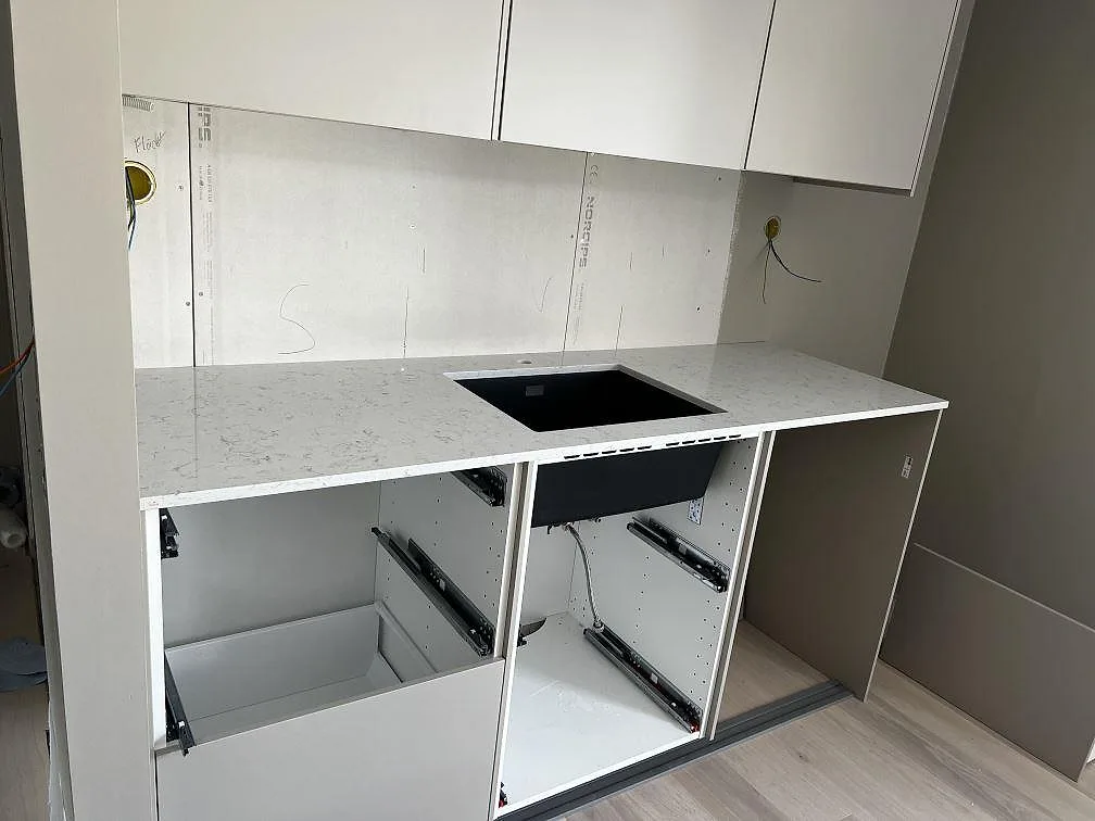 Väggbänk i polerad Technistone Noble Carrara med svart underlimmad diskho och öppna köksstommar under montering i Fjälkinge.
