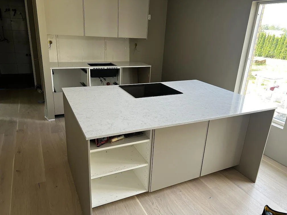 Köksbänkskiva i polerad Technistone Noble Carrara, vit komposit, på stor köksö med svart induktionshäll i Fjälkinge.