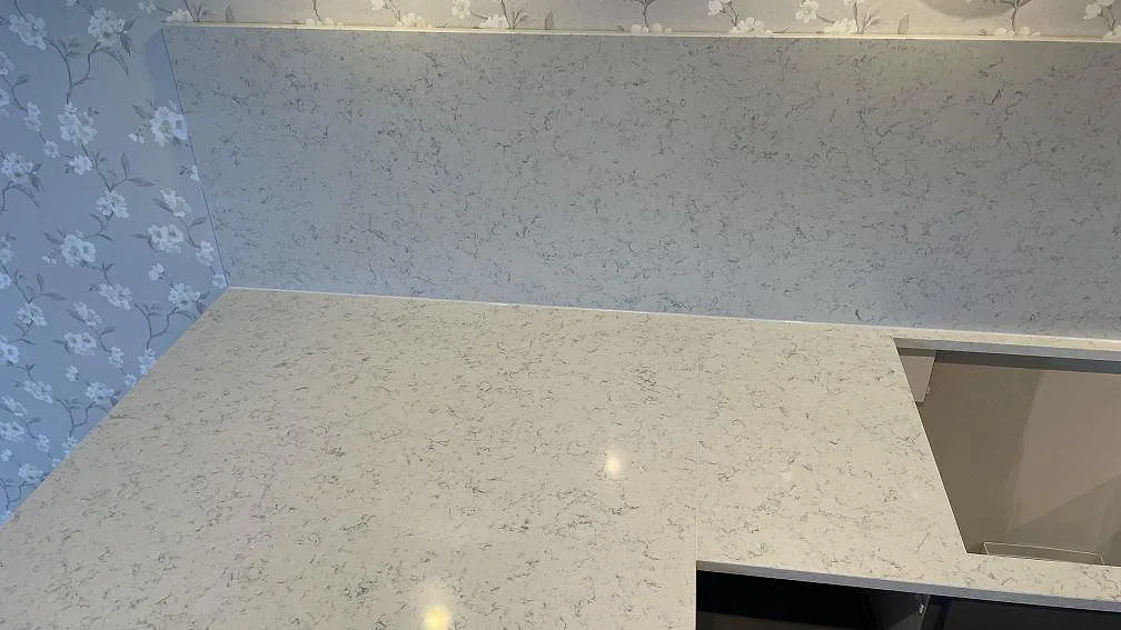 L-formad köksbänkskiva i Technistone Noble Carrara, polerad komposit, med uppvikt stänkskydd och utskärning vid diskbänken i Jönköping.