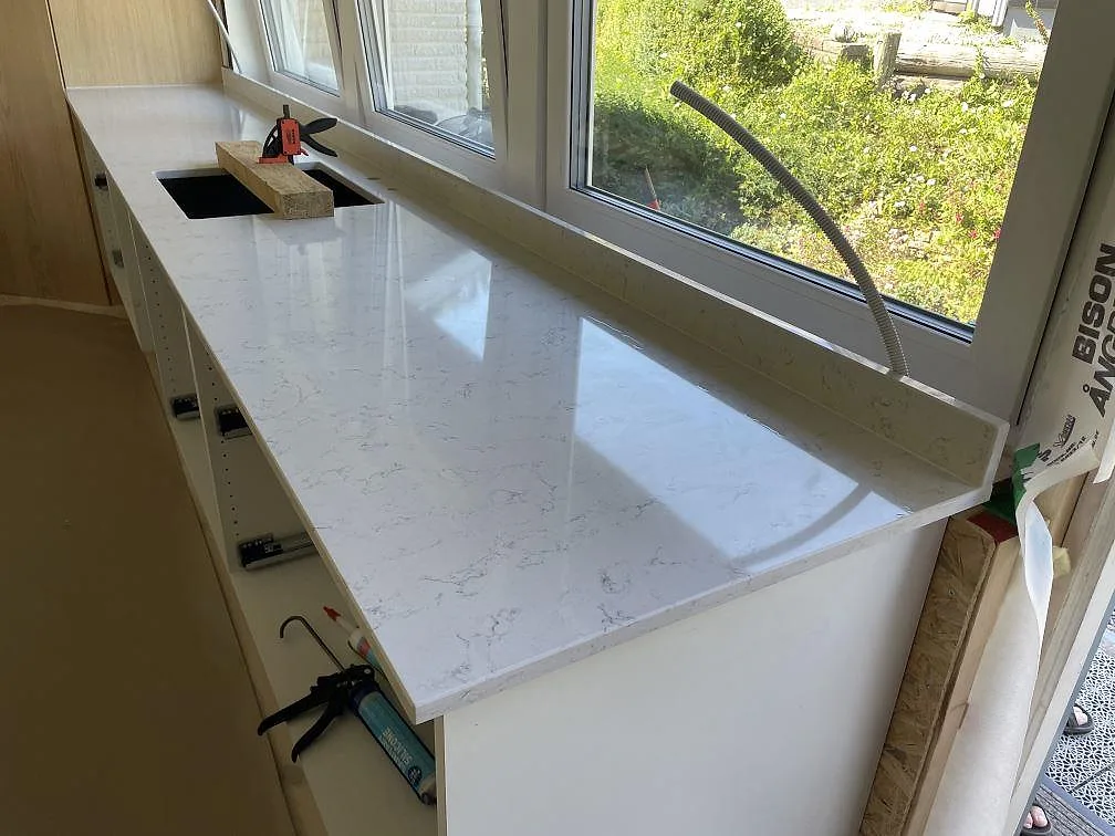 Lång fönsterbänkssida med polerad Technistone Noble Carrara, låg stänkkant och uttag för ho under montering i Lyckeby.