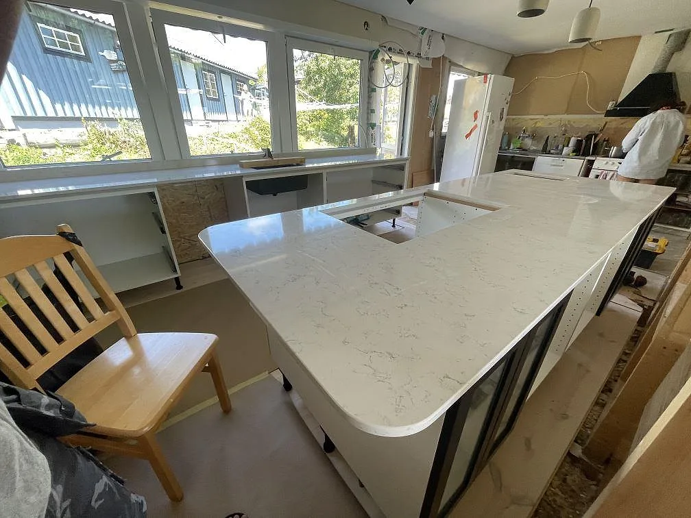 Polerad köksbänkskiva i Technistone Noble Carrara med rundat hörn, köksö och fönstersida i ett kök i Lyckeby.
