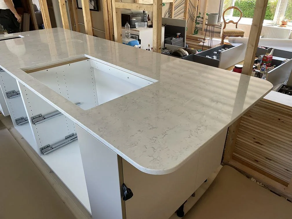 Stor köksö med polerad Technistone Noble Carrara i vit kvartskomposit och synliga cutouts under montering i Lyckeby.