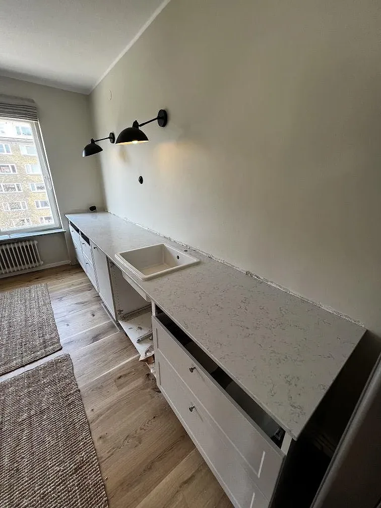 Lång köksbänkskiva i polerad Technistone Noble Carrara med underlimmad diskho längs fönstret i ett kök under montering i Malmö.