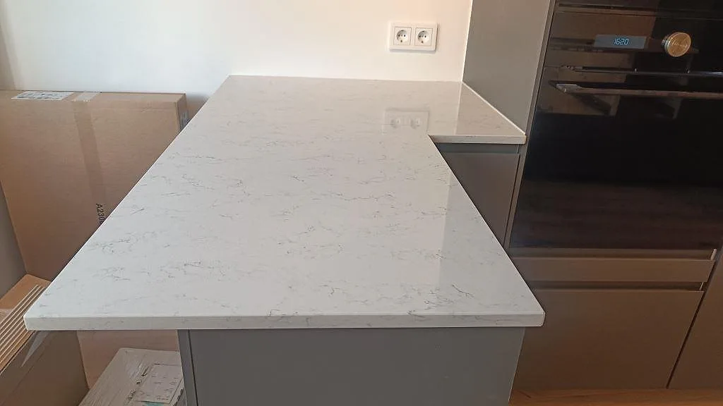 Köksbänkskiva i polerad Technistone Noble Carrara komposit, 20 mm, som halvö vid ugn och mörk träinredning i Malmö.