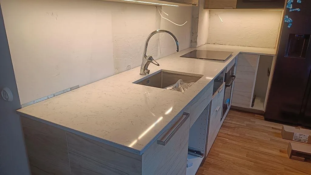 L-formad köksbänkskiva i Technistone Noble Carrara, polerad kvartskomposit, med rostfri diskho och induktionshäll i ett kök i Malmö.