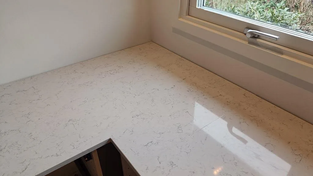 Köksbänkskiva i vit Technistone Noble Carrara längs fönstervägg, polerad kvartskomposit med grå ådring i ett kök i Staffanstorp.