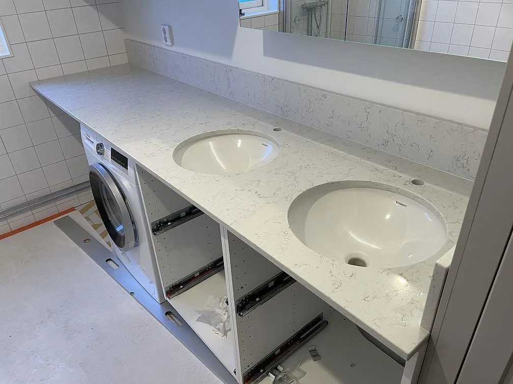 Badrumsbänkskiva i vit Technistone Noble Carrara, polerad kvartskomposit, med två underlimmade tvättställ och uppvikt stänkkant i Uppsala.