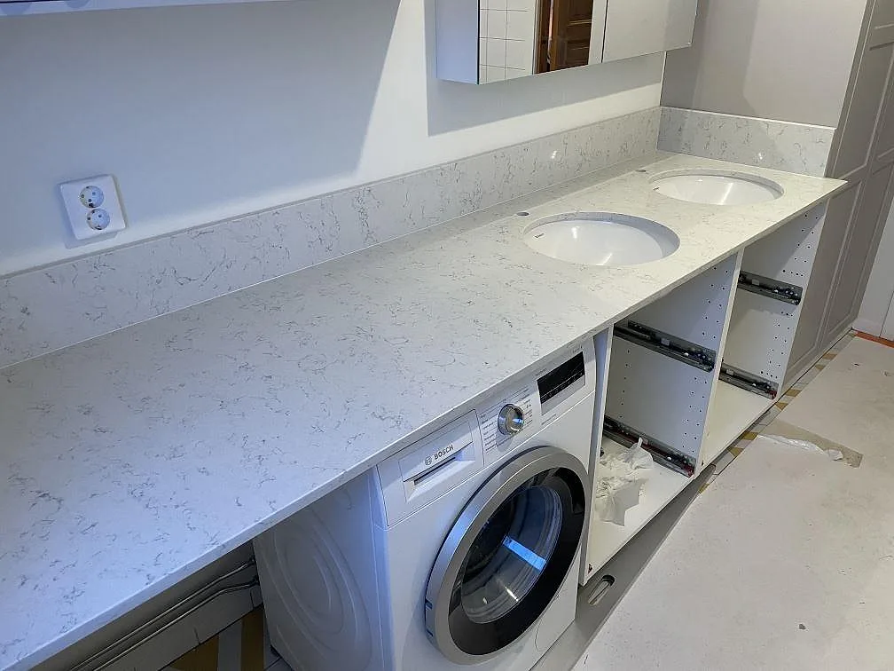 Polerad Technistone Noble Carrara som badrumsbänkskiva med dubbla underlimmade tvättställ och tvättmaskin under skivan i Uppsala.
