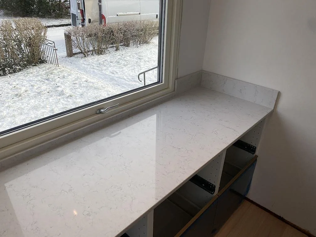 Ljus kompositskiva i Technistone Noble Carrara, polerad, som arbetsyta vid fönster med blågrå köksstommar i Värnamo.