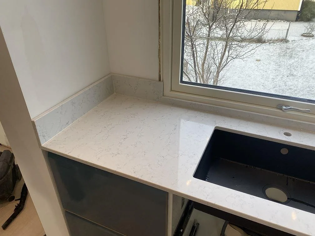 Närbild på polerad Technistone Noble Carrara som köksbänkskiva med svart diskho och fönsterljus i Värnamo.