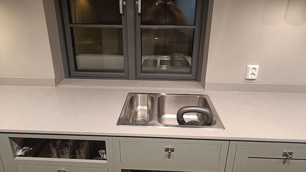 Diskbänksdel med polerad Technistone Noble Concrete Grey kvartskomposit i Bankeryd, låg stänklist och rostfri dubbelho framför fönster.