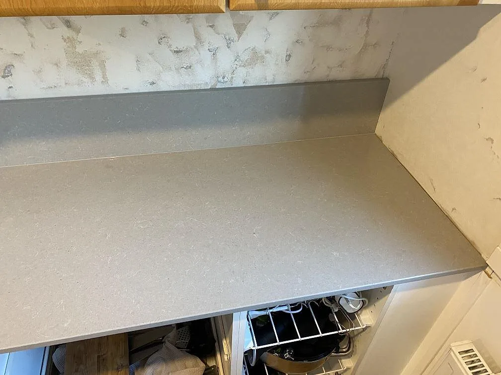 Höger del av köksbänkskiva i polerad grå Technistone Noble Concrete Grey med låg stänkkant och öppen stomme under montering i Beddingestrand.