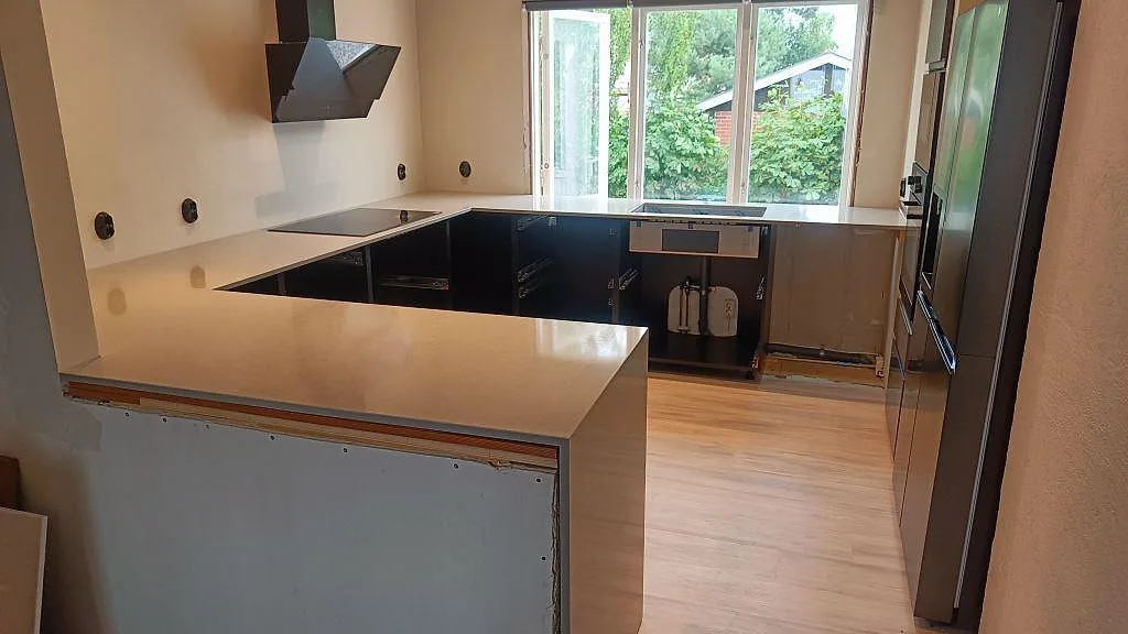 Köksbänkskiva i Technistone Noble Concrete Grey, polerad komposit, i ett U-format kök med halvö, induktionshäll och stort fönsterparti i Höllviken.