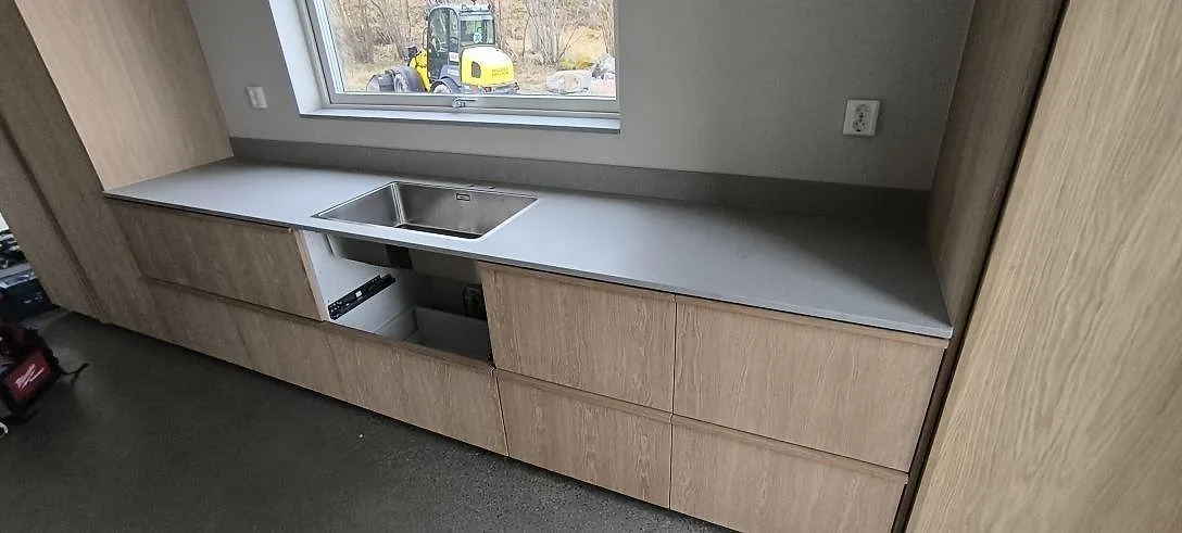 Matt köksbänkskiva i grå Technistone Noble Concrete Grey, kvartskomposit, längs fönstervägg med diskhoområde och stänkskydd i ett kök i Ingarö.