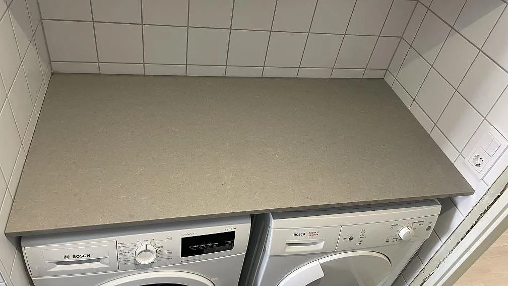 Bänkskiva i grå Technistone Noble Concrete Grey, polerad kvartskomposit, över två tvättmaskiner i ett kaklat tvättutrymme i Malmö.