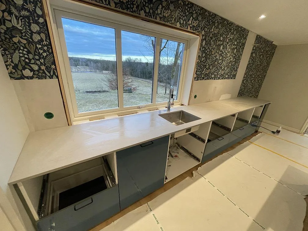 Bänkrad i grå Technistone Noble Concrete Grey längs fönstret med underlimmad rostfri diskho och blandare i ett kök i Värnamo med utsikt mot landskapet.