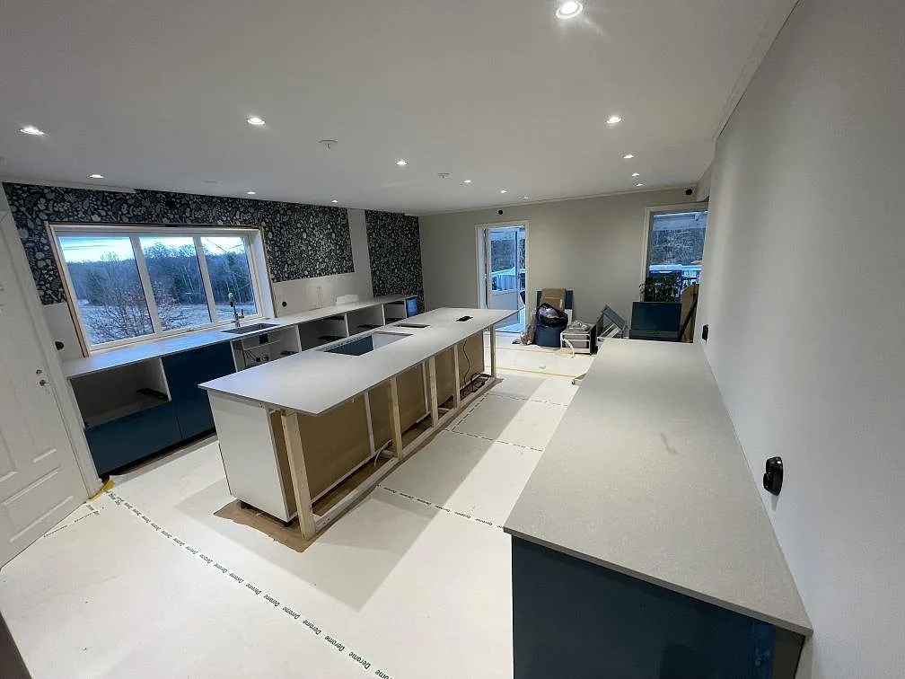 Översikt av kök i Värnamo med köksö och bänkrad i matt grå Technistone Noble Concrete Grey, öppna stommar och blommig tapet under montering.