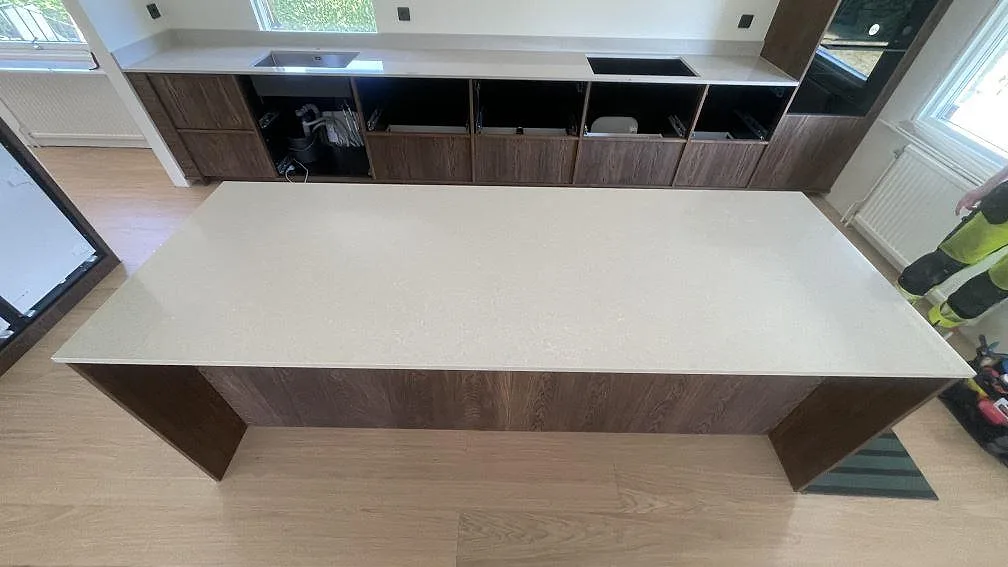 Översikt över en fri köksö och väggbänk i polerad Technistone Noble Ivory White med låg stänkkant och mörka trästommar i Åhus.