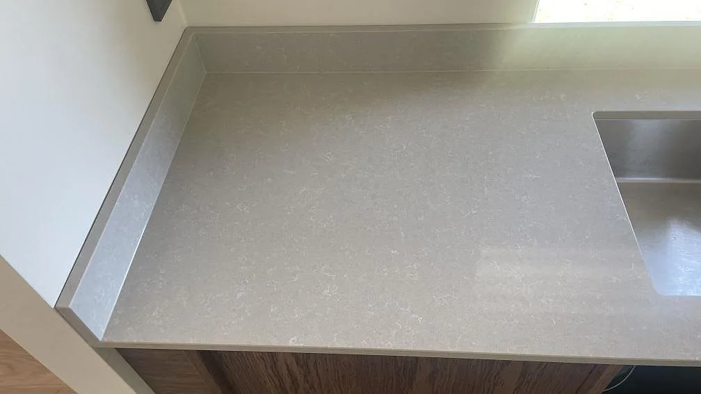 Hörn av köksbänk i polerad Technistone Noble Ivory White med låg uppvikt stänkkant och rostfri diskho vid fönster i Åhus.