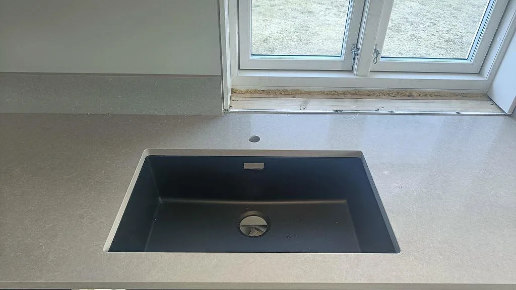 Underlimmad svart kompositdiskho i en polerad bänkskiva i Technistone Noble Ivory White, med separat borrat blandarhål, i ett kök i Fritsla.